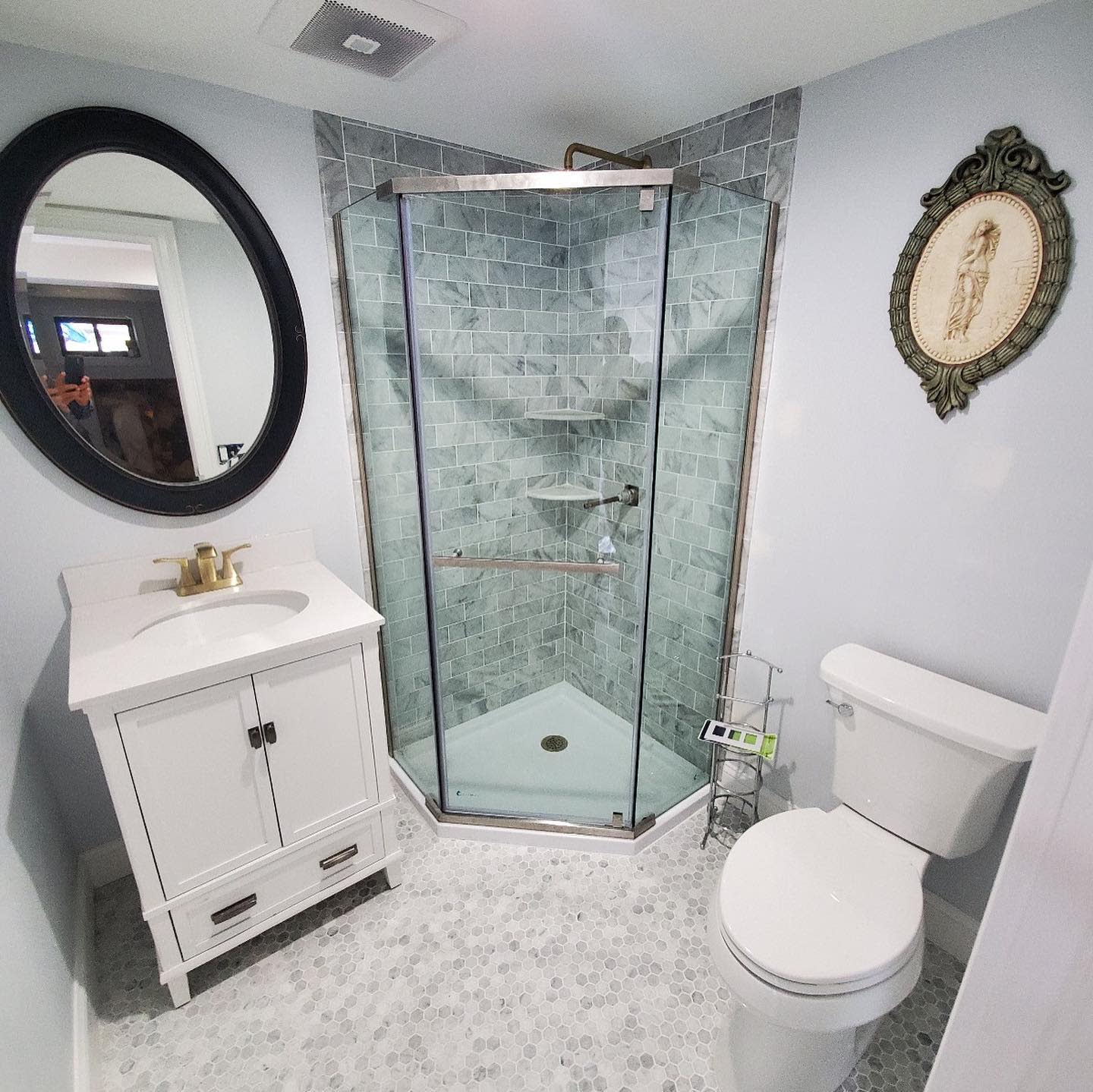 White bathroom with corner glass shower, vanity sink, round mirror, and decorative wall art.